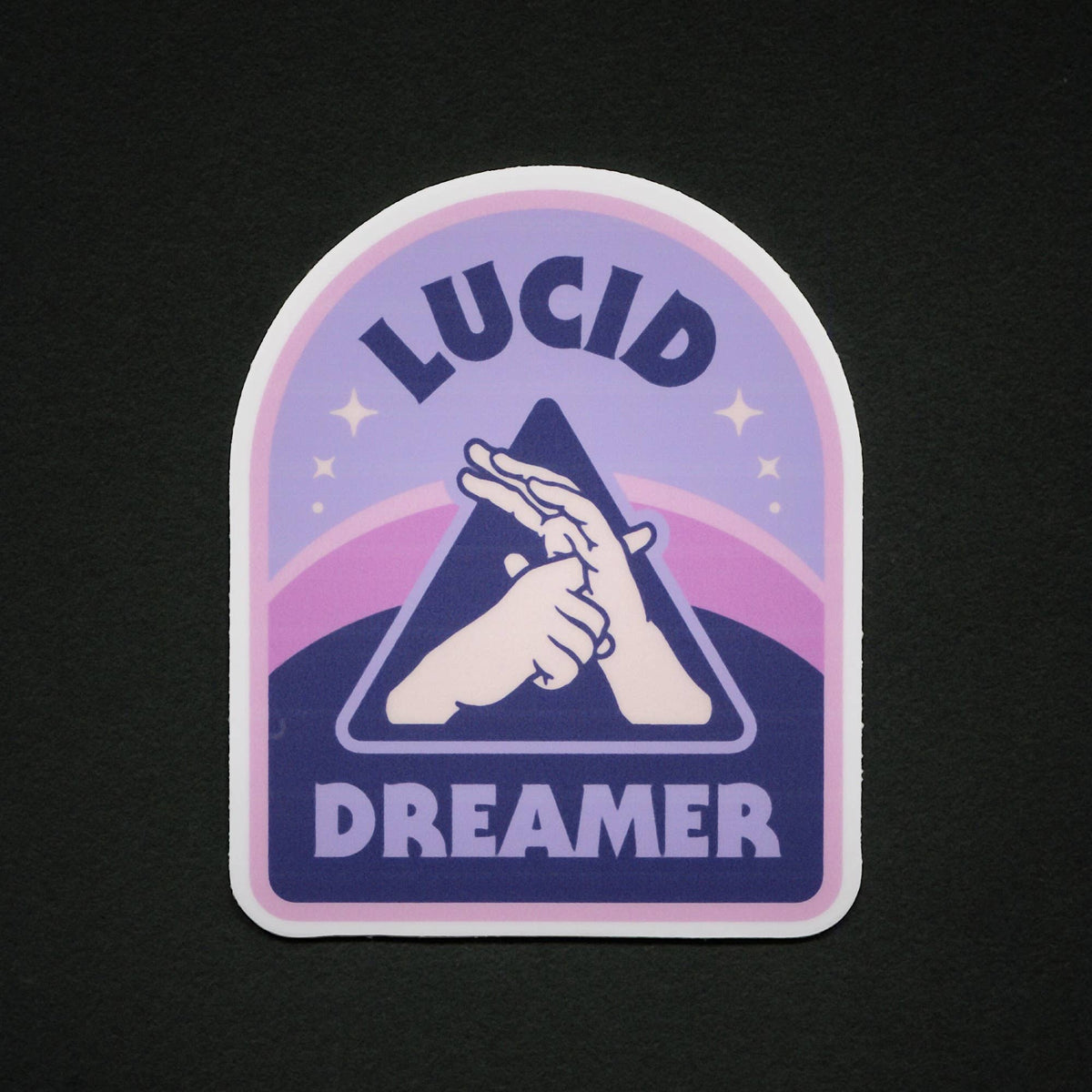 A die-cut sticker with an arched top and rounded bottom edge, featuring the words 'LUCID DREAMER' in dark purple lettering around a central graphic of two hands forming a star. The background of the sticker has a purple-to-pink gradient with small star accents, all set against a solid black background.
