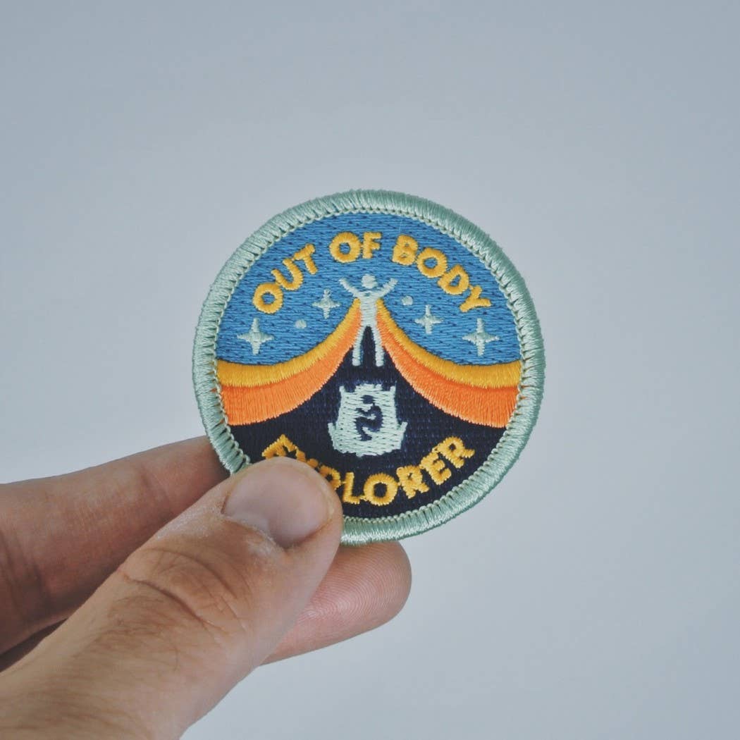 A hand holding a circular embroidered patch that reads 'OUT OF BODY EXPLORER' with a blue, orange, black, and light green design on a plain light gray background.