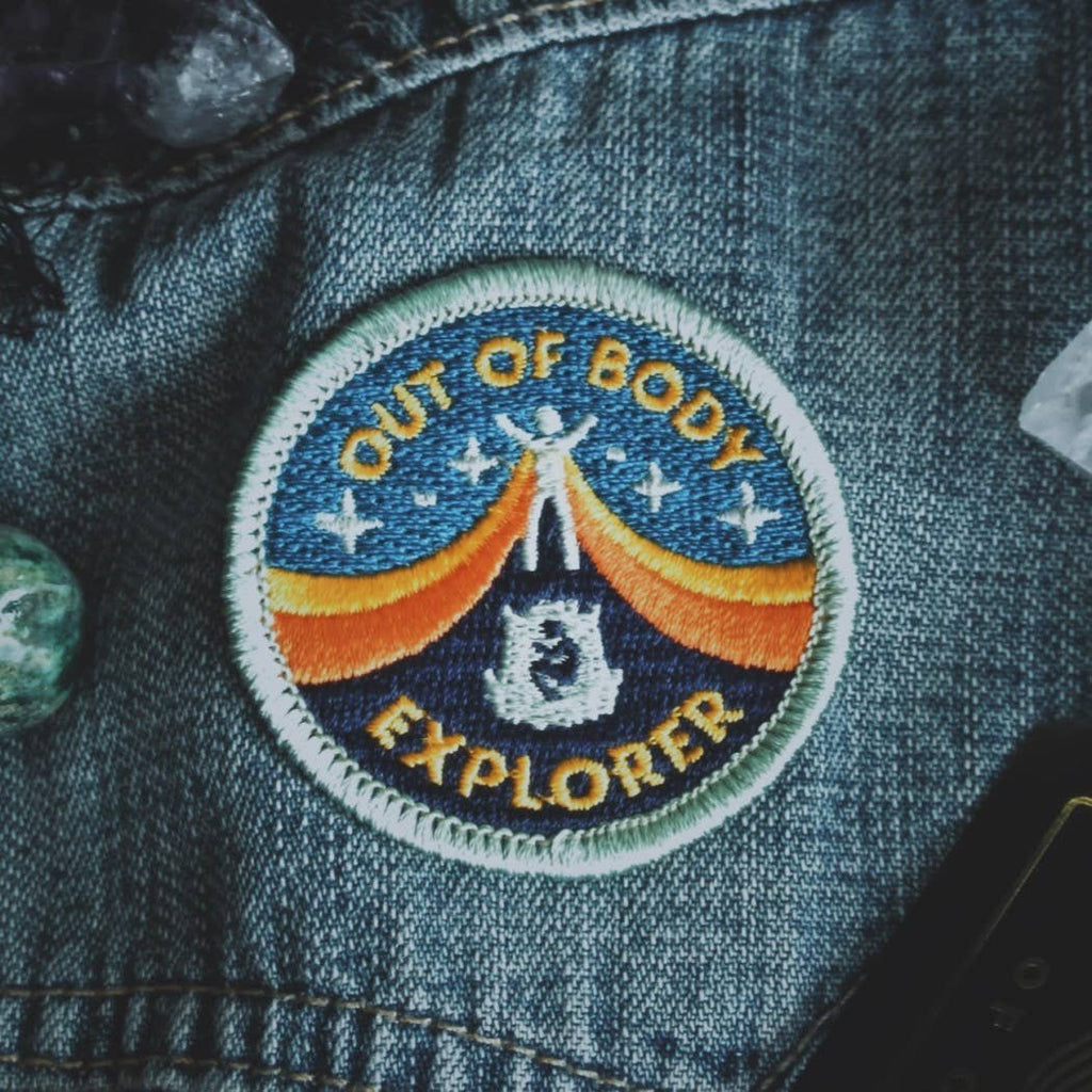 A circular embroidered patch reading 'OUT OF BODY EXPLORER' with a blue sky, orange arc, and white astronaut design, attached to dark blue denim fabric.