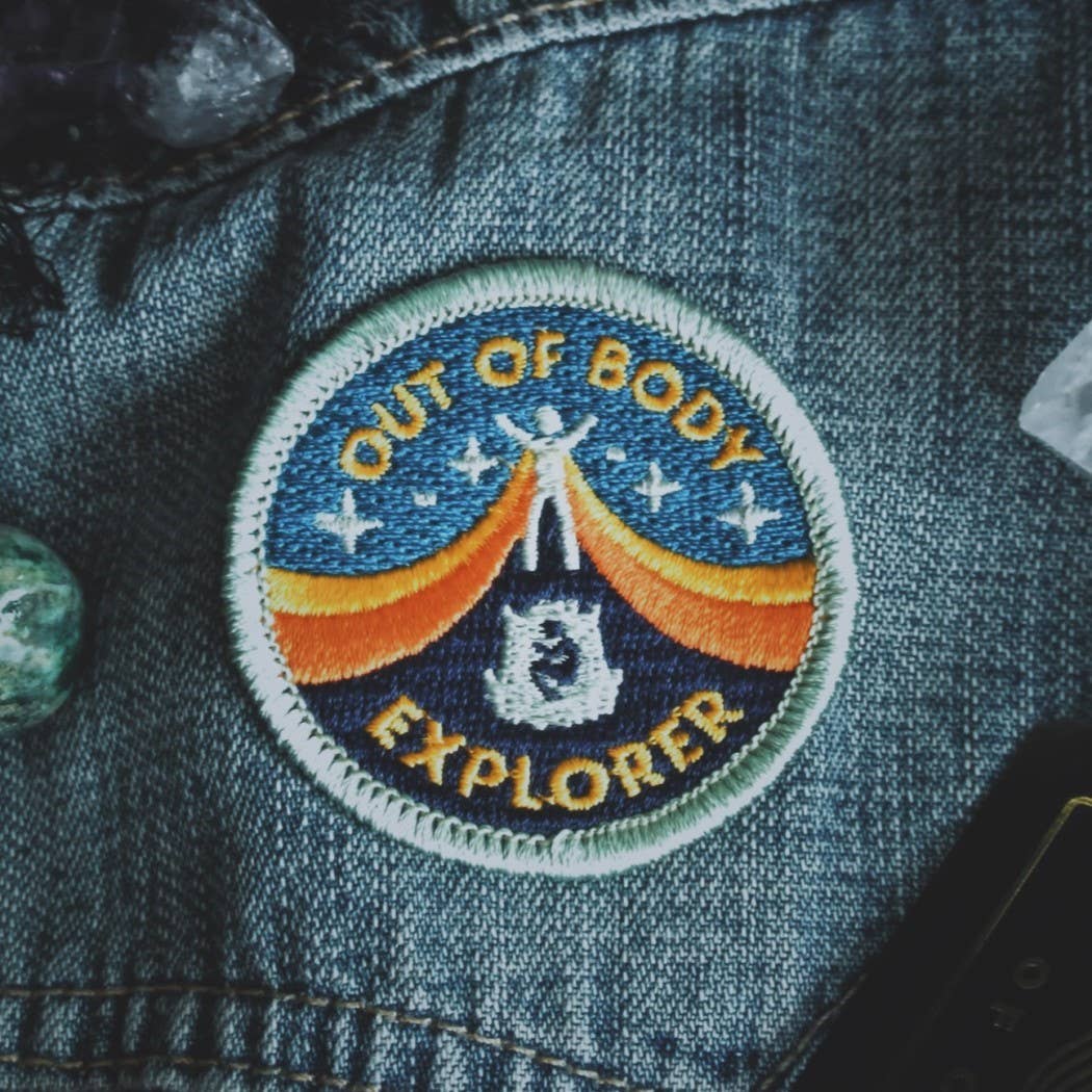 A circular embroidered patch reading 'OUT OF BODY EXPLORER' with a blue sky, orange arc, and white astronaut design, attached to dark blue denim fabric.