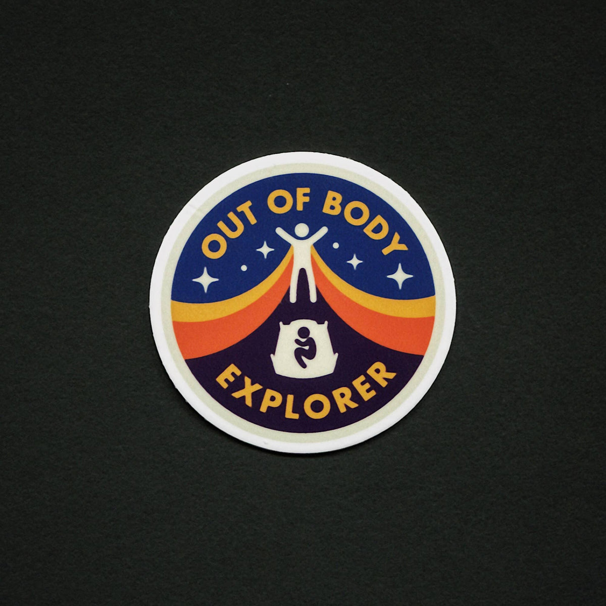 A circular sticker with the words 'OUT OF BODY EXPLORER' in yellow around a central graphic of a white figure standing on a rainbow arch over a dark blue and purple landscape, shown on a black background.